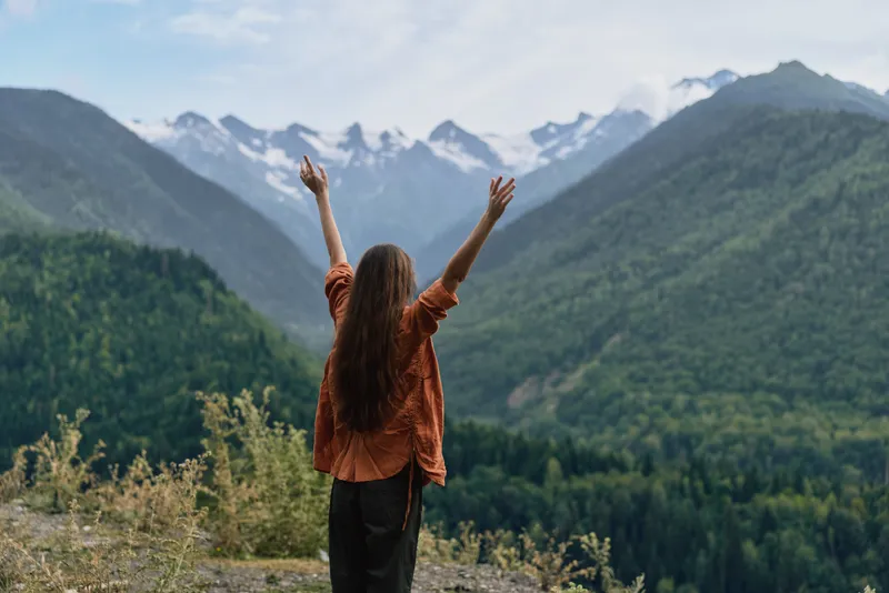 Woman stands on a hillside with arms raised toward snowcapped mountains, expansive valley below, bright sky, capturing a moment of freedom, adventure and natural inspiration and wonder.
