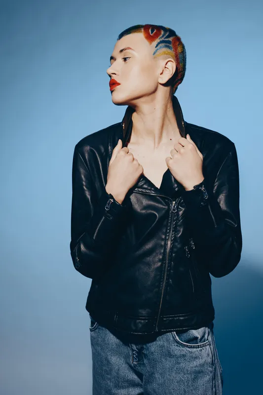 edgy woman with colorful short hair wearing a black leather jacket and casual jeans against a blue background, stylish urban fashion portrait, contemporary look