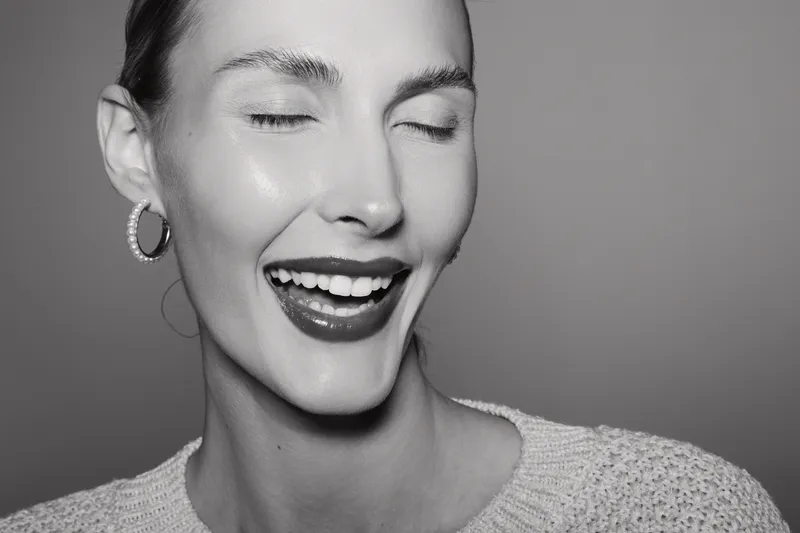 Close-up of a smiling caucasian woman with fair skin and earrings, eyes closed, expressing joy in soft studio lighting on a grey background.