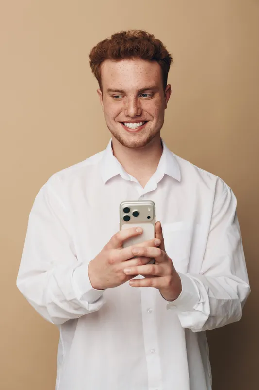 Smiling young man with red hair wearing white shirt looks at smartphone with happy expression. Isolated on beige background. Concept of technology, communication, and positive emotions.