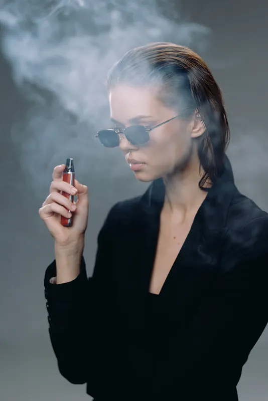 Woman vaping in black outfit with sunglasses, producing smoke in moody lighting environment.