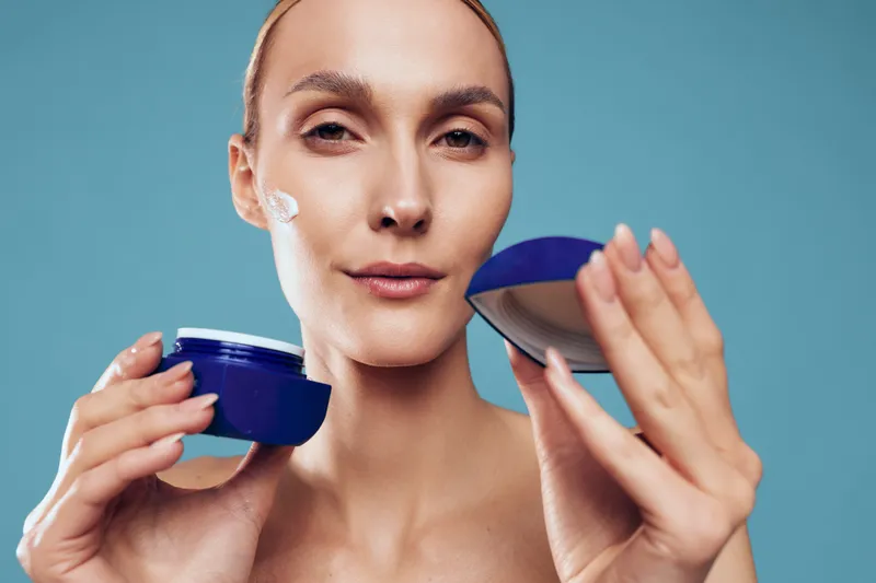 Young woman applying face cream on clean skin holding an open blue jar with soft natural makeup and smooth skin against light blue background closeup beauty concept