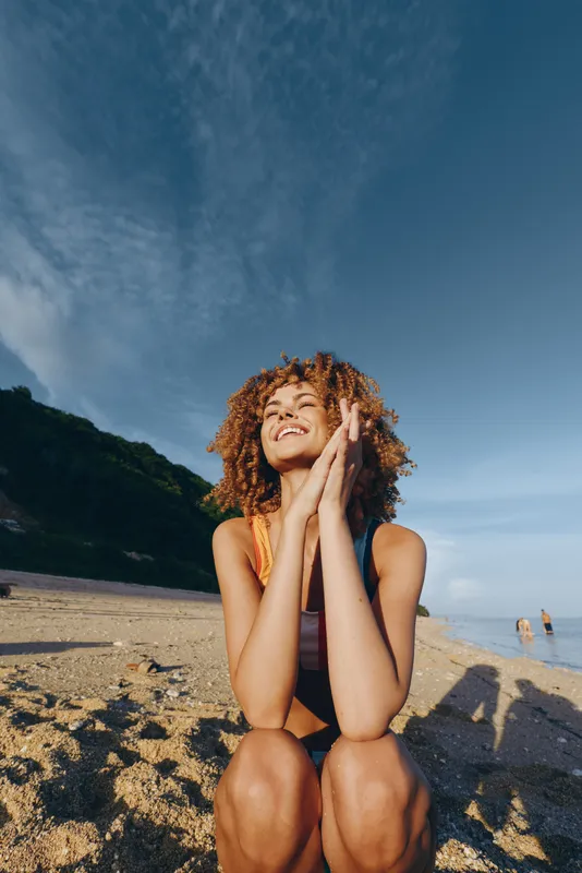 A relaxed woman on the beach, curly hair, smiling in summer sunshine, outdoor lifestyle concept, calm relaxation on a sandy shore, warm summer escape for vacation and travel memories today