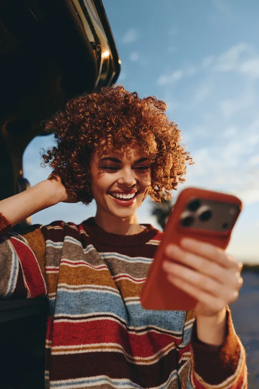 Woman lifestyle with smartphone smiling curly hair joy near a car window outdoors in sunny daylight featuring relaxed laughing communication in a casual striped sweater for travel