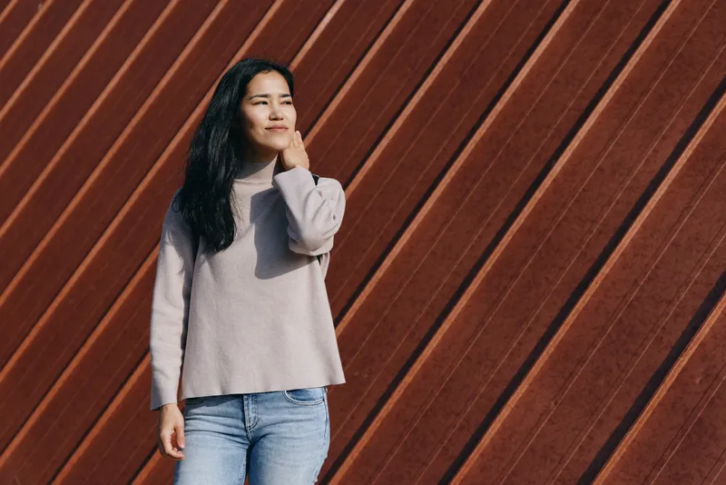 Woman in a cozy knit sweater and denim stands against diagonal wooden panels, calm expression, natural light, casual fashion, urban backdrop, soft textures