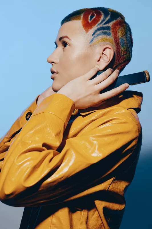 Woman with colorful shaved hairstyle wearing a yellow jacket, looking sideways thoughtfully against a blue background in a modern portrait style.