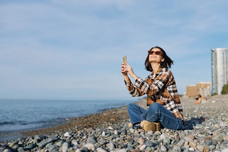 Woman sits on a pebbled beach by the sea, wearing a plaid shirt and jeans, taking a selfie with a smartphone, bright sky and distant shoreline in the background