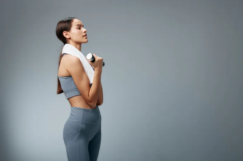 Young woman fitness portrait lifting a small dumbbell in a grey athletic set with a white towel draped over her shoulder, side profile in a minimalist studio background, focused training concept.