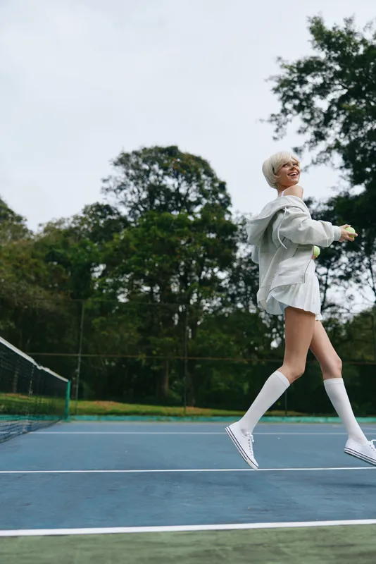 Young female model playing tennis on a sunny day, wearing a casual outfit including a white oversized shirt and knee high socks, exuding joy and energy