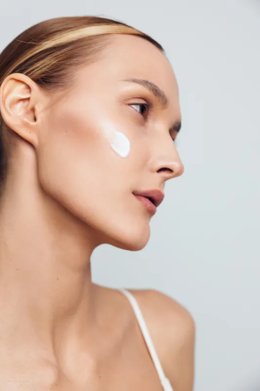 Close profile of a young woman applying sunscreen on her cheek in a studio with soft lighting, calm expression, and natural skin glow.