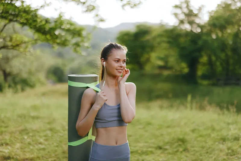 Young woman enjoying outdoor yoga practice in green forest, carrying yoga mat, wearing comfortable athletic attire, and smiling, embodying wellness and fitness lifestyle
