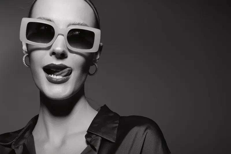 Close-up of a pale white woman with slicked hair wearing oversized sunglasses and earrings, tongue out playfully. Fashion concept with attitude and monochrome studio lighting.