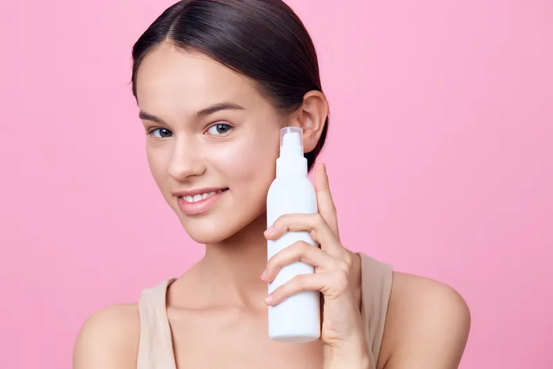 Young woman holding a skincare product against a pink background, exhibiting a joyful expression, embodying beauty and self care