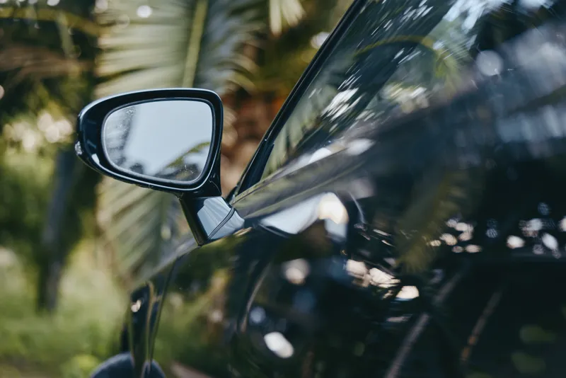 car side mirror glossy black with palm reflection, tropical outdoor closeup showing vehicle detail and faint young adult driver reflection in casual outfit, travel lifestyle vibe
