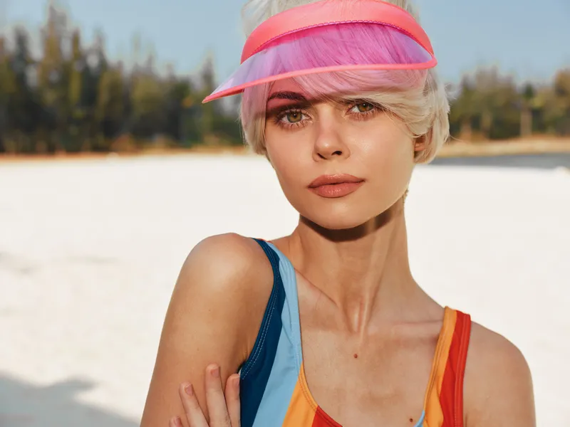 Closeup portrait of a stylish woman with short pink hair wearing a pink visor and colorful swimsuit on a sunny beach, sunlit skin and confident gaze.