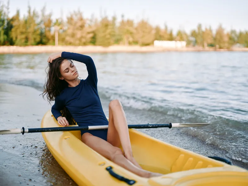 Female kayaker wearing a navy long sleeve top sits in a yellow kayak near a calm shoreline, holding a paddle across her lap beneath clear sunny skies.