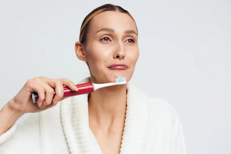 A woman in a white bathrobe holds a red toothbrush near her face with a skeptical expression. The plain background emphasizes her doubtful mood and personal care moment.