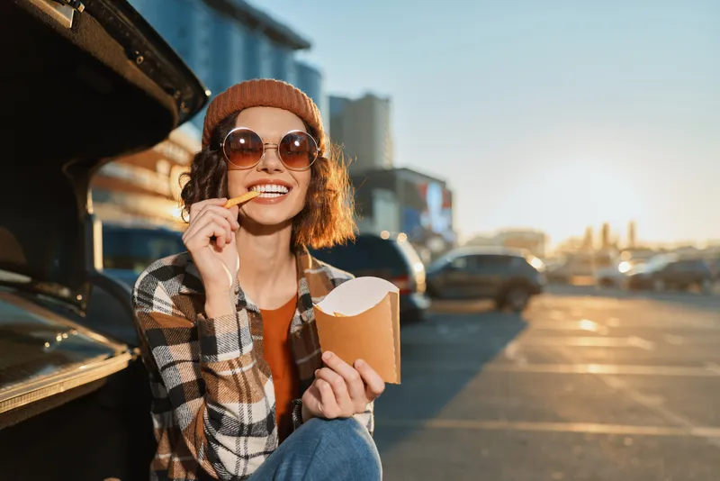 Woman eating fries by car in parking lot, smiling in sunglasses and beanie during golden hour glow. Authenticity, candid lifestyle and mindful living captured in emotional storytelling.