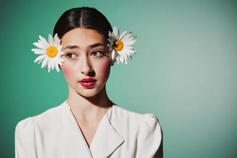 Young woman with two daisies in her braided hair and natural makeup wearing a white blouse on green background. Creative fashion and beauty concept with floral details.