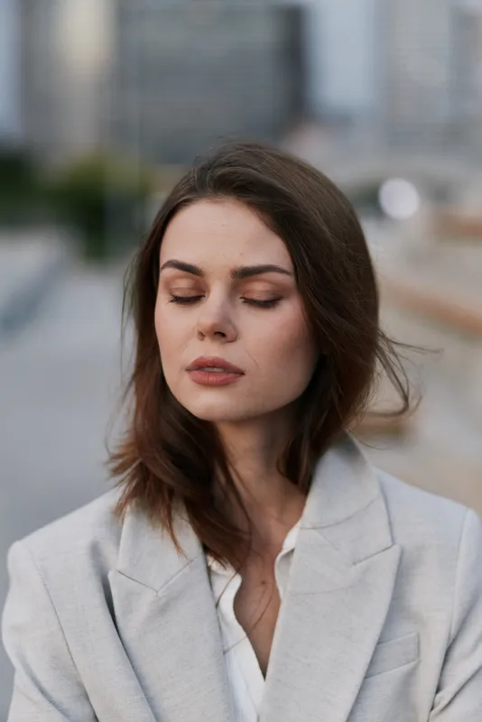 Confident woman in a business suit with closed eyes, embracing calmness against an urban backdrop, exuding professionalism and poise in a modern city setting.