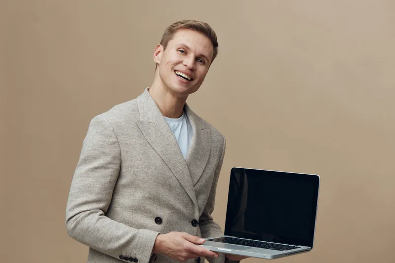 smiling man holding laptop wearing grey blazer digital nomad remote work concept beige background cheerful happy young adult