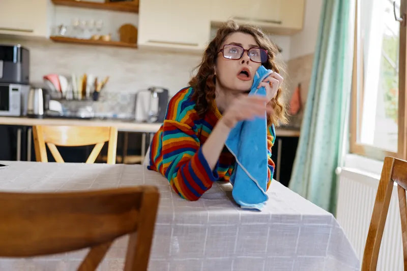 A woman sits at a dining table in a home kitchen, talking on a phone and looking surprised, wearing a colorful striped sweater and glasses, capturing a casual home moment.