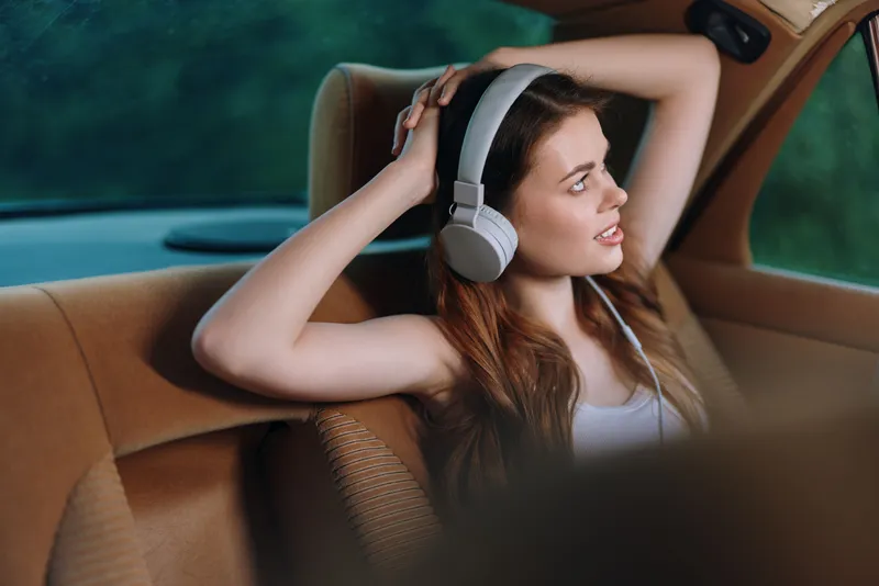 Young woman with long hair enjoying music in a car, wearing headphones, relaxed, emotional expression, sunny day, natural background.