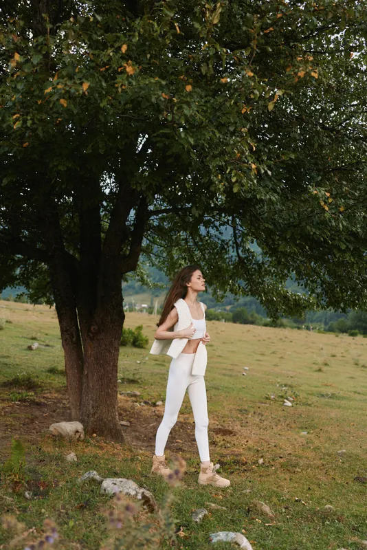 Cheerful woman outdoors, in white outfit, strolling near a large tree, sunlit field moment with relaxed posture and light breeze, natural colors and serene mood