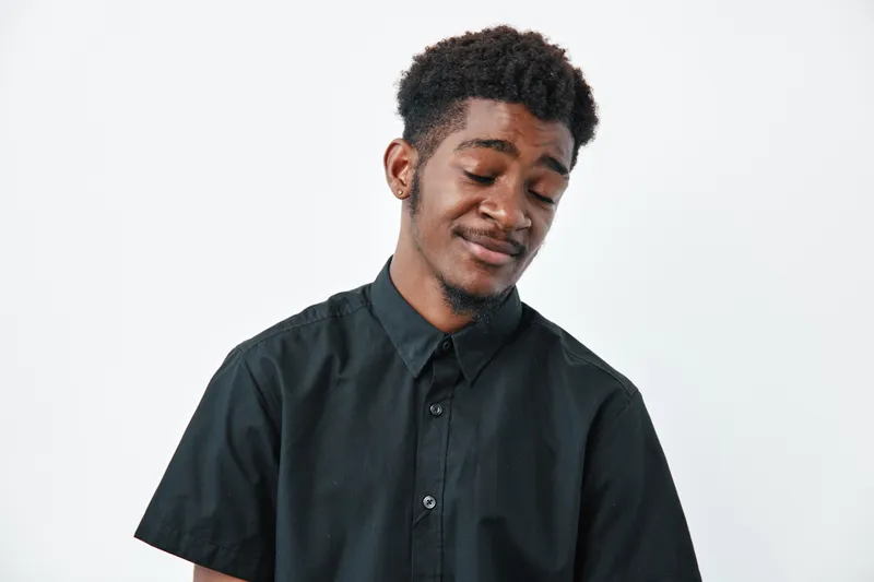 Young African American man with dark skin and curly hair wearing black shirt standing with eyes closed and slight smile on white background in studio portrait.