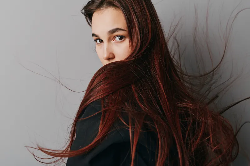 Young woman with long flowing hair looks over shoulder against plain background, wearing dark clothing with natural makeup and serious expression in studio portrait.