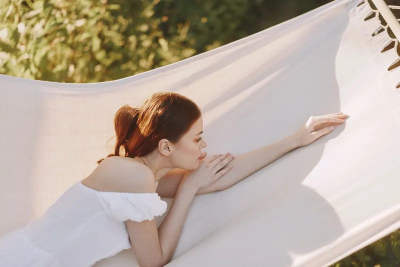 Young woman relaxing in a hammock during summer, wearing a white dress, enjoying a peaceful moment in a sunny green garden, reflecting tranquility and serenity