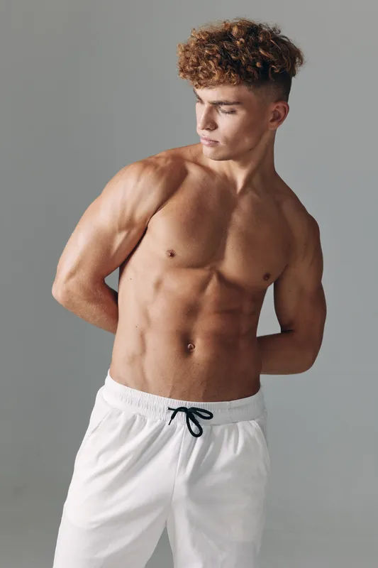 Young muscular man standing shirtless with white pants on gray background showing athletic body and defined muscles with curly hair and pleasant facial expression