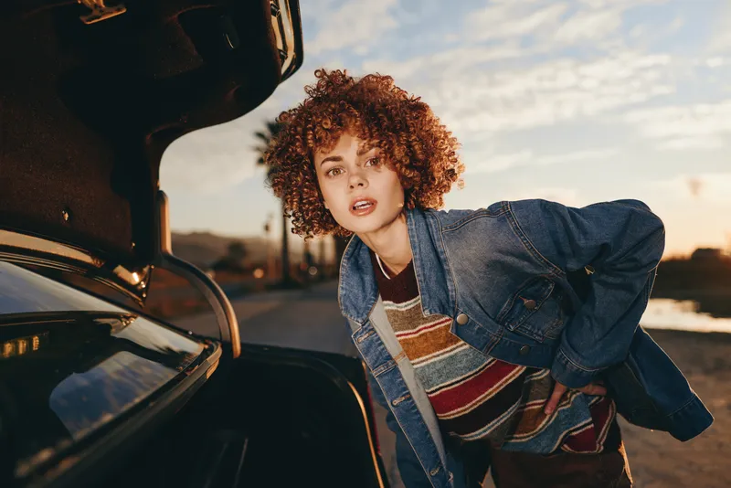 Woman curly hair denim jacket leaning at open car hood during sunset, lifestyle moment on roadside, casual street fashion, soft warm light, checking engine needs