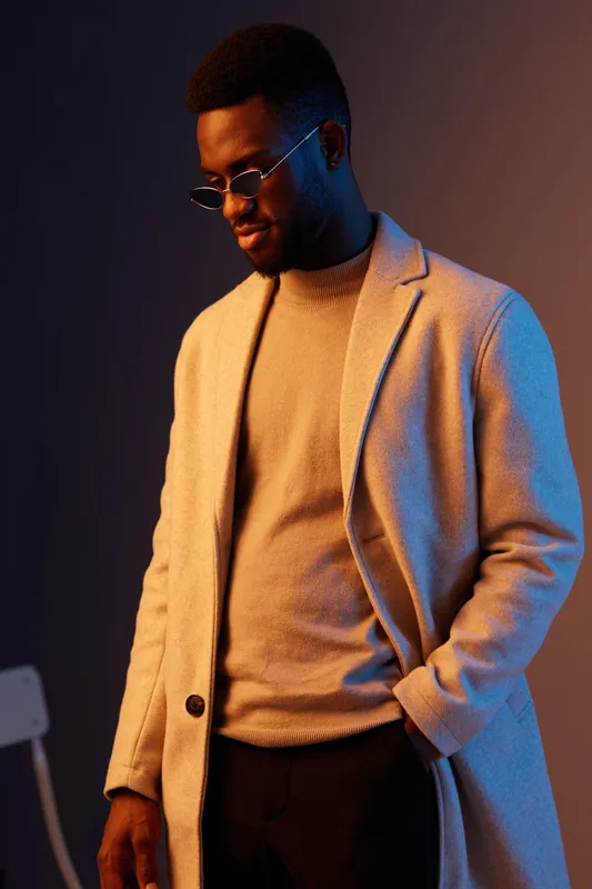 Stylish young African man in a beige coat and sweater, showcasing confidence and elegance against a moody blue background, perfect for fashion campaigns and lifestyle imagery