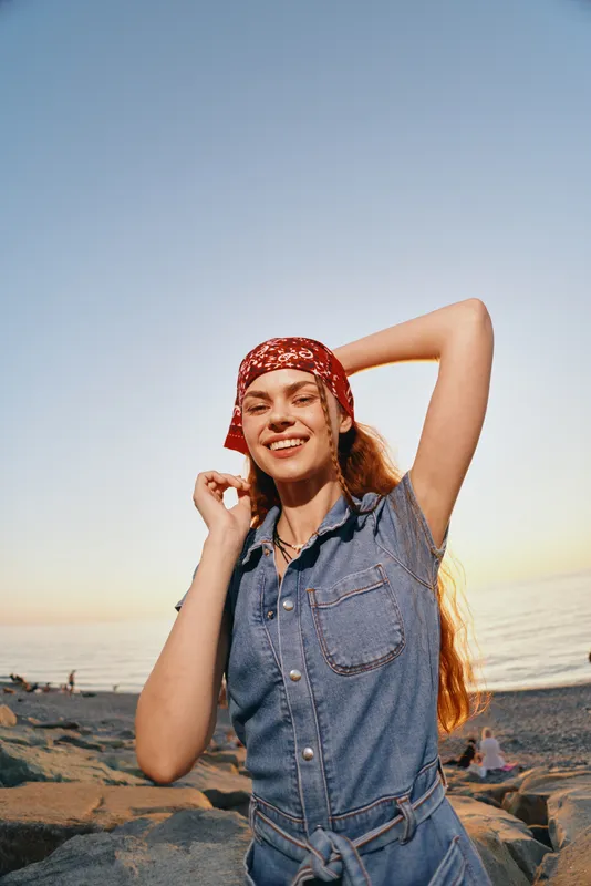 smiling woman wearing red bandana and denim dress enjoying sunset at beach with calm sea and clear sky background