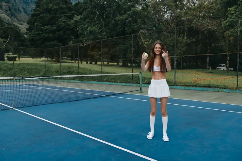 Young female tennis player celebrating victory on the court, wearing a white sports outfit, with a joyful expression, surrounded by green trees in a vibrant setting
