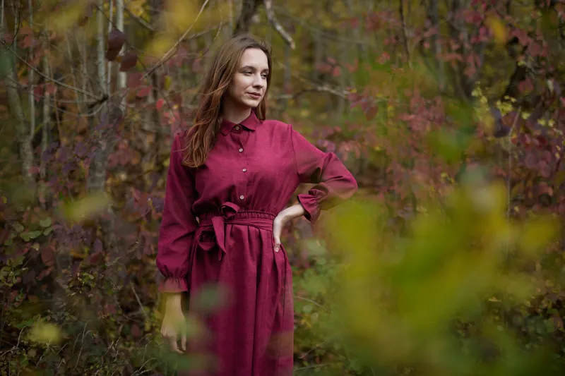 A young woman in a burgundy dress stands among autumn shrubs, posing naturally with a calm confident gaze; a genuine, realistic portrait that emphasizes a true-to-life outdoor character and mood.