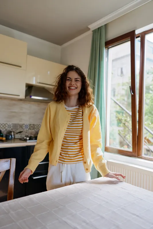 Smiling woman in a bright kitchen wearing a yellow cardigan and striped t shirt, leaning on a table. Casual home lifestyle scene emphasizing warmth, comfort, and everyday joy.