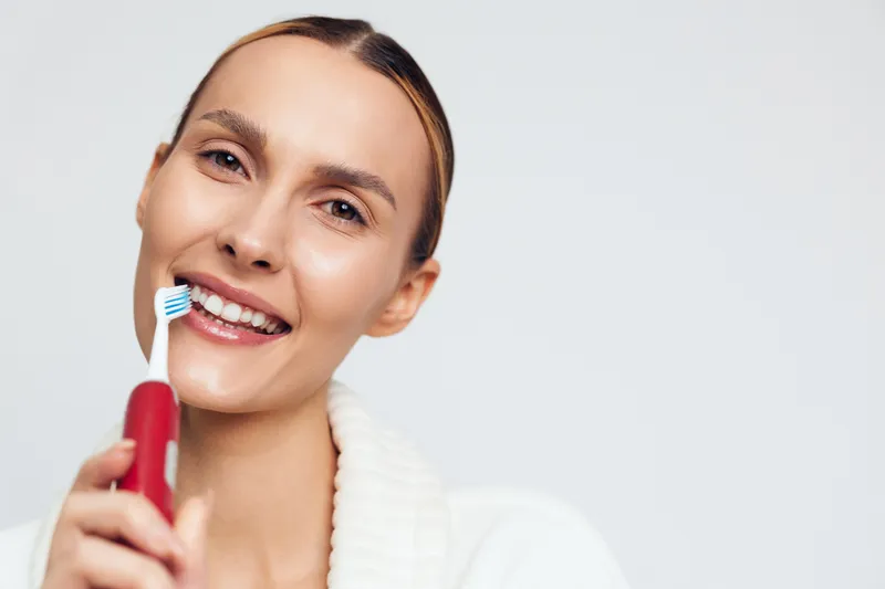 Smiling woman holding electric toothbrush close to face, clean teeth visible, fresh skin, white background, healthy lifestyle, dental hygiene concept, natural light