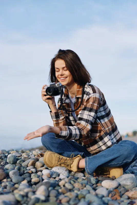 Woman photographer on a rocky beach, kneeling with a camera, wearing plaid shirt and boots, capturing candid moments by the shoreline under a clear sky, and sunlight