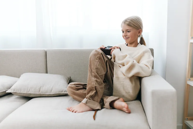Authentic scene of a student girl at home, relaxing on a sofa with a game controller, capturing lifestyle, play, and school related moments in a natural setting for audiences seeking real everyday