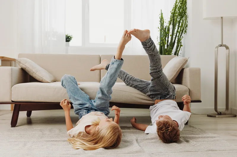 Two children lie on the floor playing with feet raised, enjoying playful moment in bright living room with modern sofa and natural light. Concept for childhood fun and family time.