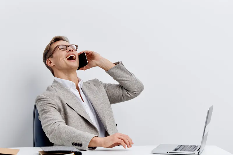 laughing man talking smartphone gray blazer white shirt glasses sitting at desk with laptop working remote digital nomad concept