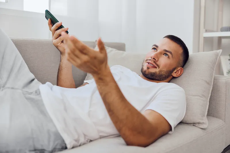 Relaxed man enjoying a phone call, expressing emotions while lounging on a sofa, reflecting modern challenges of work-life balance and emotional vulnerability.