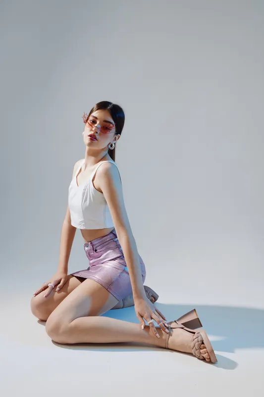 Fashionable woman in a white top and shiny skirt poses on the floor wearing stylish sunglasses and high heel sandals. Studio shot on a neutral background. Concept of beauty and fashion.