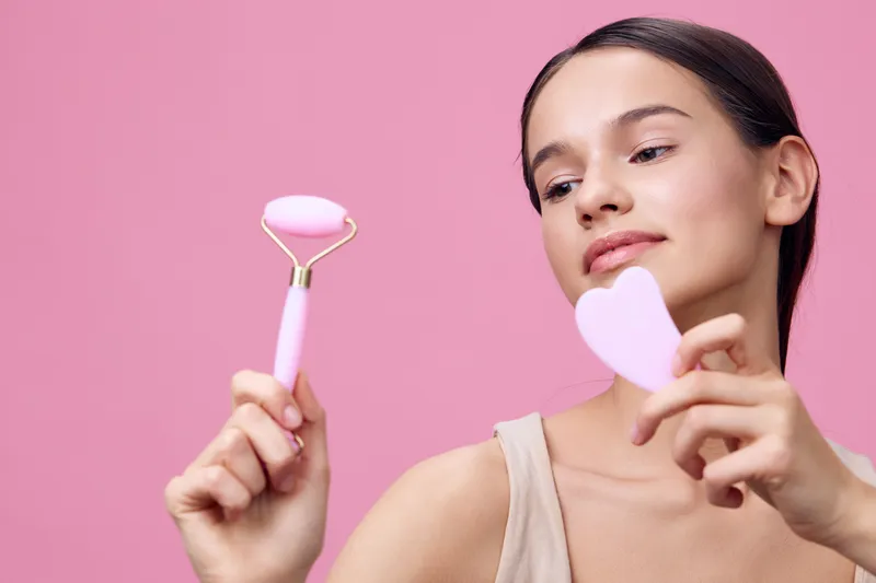 Young woman showcasing skincare tools, a facial roller and a heart shaped gua sha on a pastel pink background, emphasizing beauty and self care