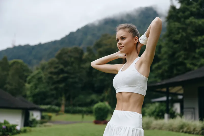 Athletic young woman in a white sports outfit stretching outdoors with green mountains in the background, embodying health, fitness, and well being
