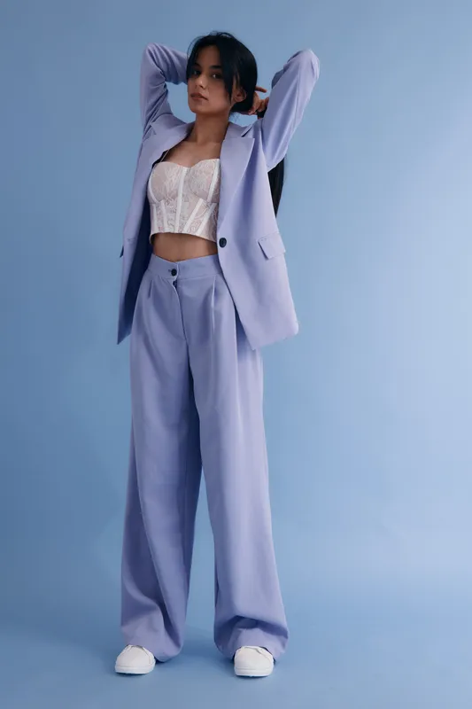 Young woman in light purple suit and white corset posing with arms raised against a blue background, showcasing modern fashion and casual style.