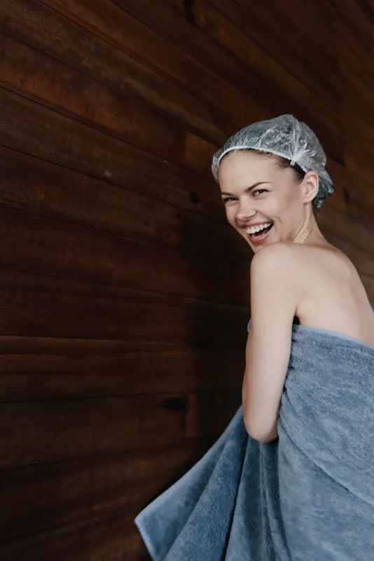 smiling woman wrapped in towel with shower cap laughing by wooden wall, natural beauty, spa wellness, carefree and joyful moment, cozy indoor atmosphere