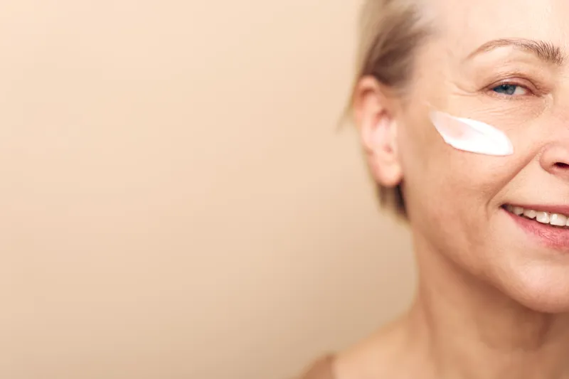 Smiling mature woman with short hair applies white cream on cheek, close-up on beige background. Concept of skincare, antiaging, selfcare, and facial beauty for older adults.